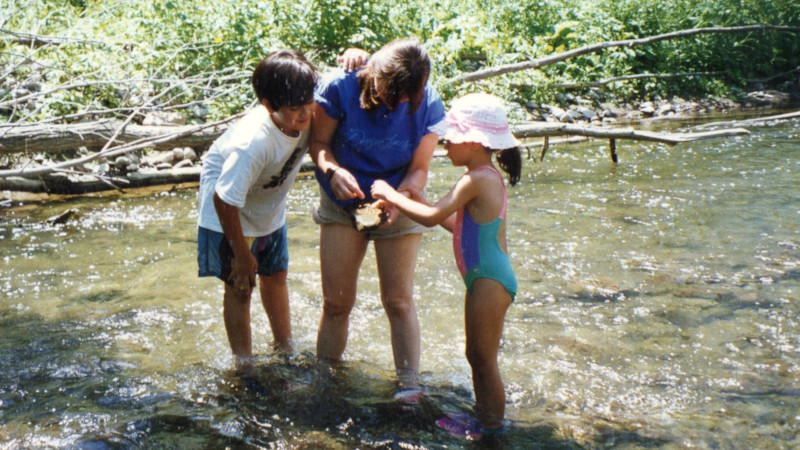 Photo of a 10 year-old boy, a middle aged woman, and a 6 year-old girl standing in a creek and all looking at a rock, with the woman pointing to it.