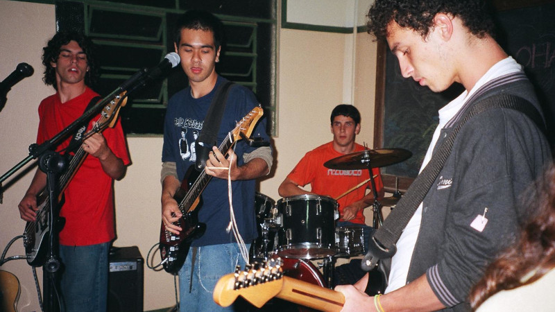Photo of a guy with curly hair playing the base guitar, me (half-asian guy with short hair playing guitar), a drummer in th background, and another guitar player with brown curly hair.