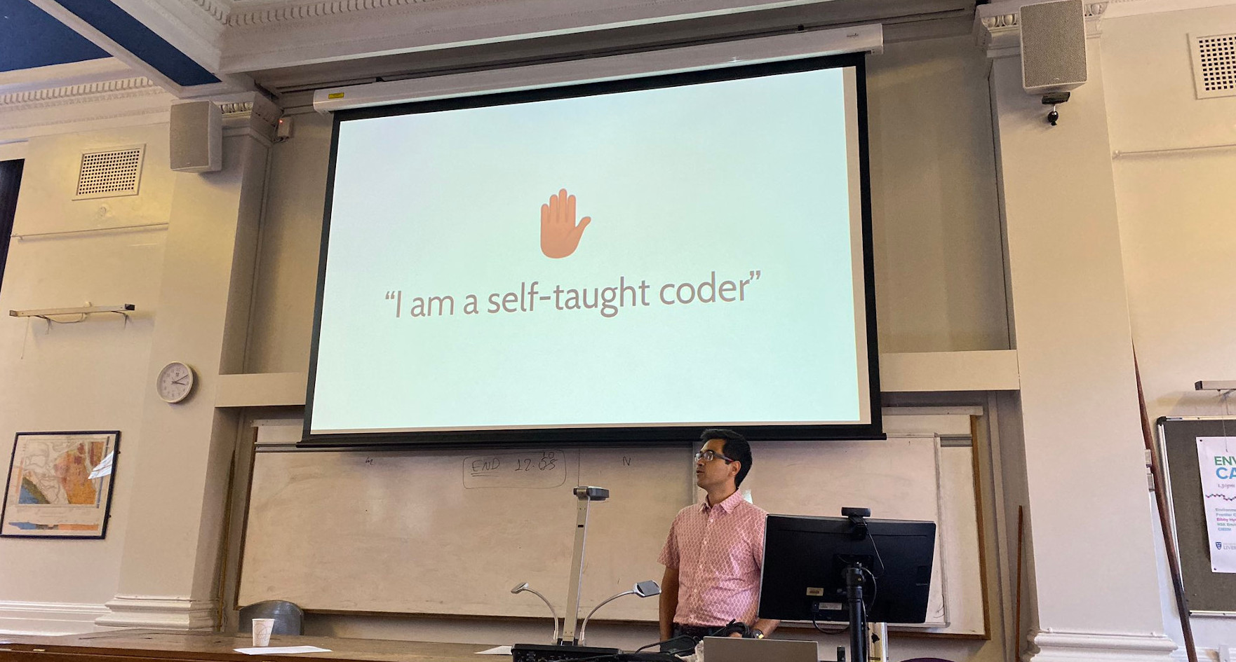 Photo with a projection screen at the center (slide has a hands-up emoji and the phrase 'I am a self-taugh coder') and a slightly tan man in a red shirt standing in front of the screen.