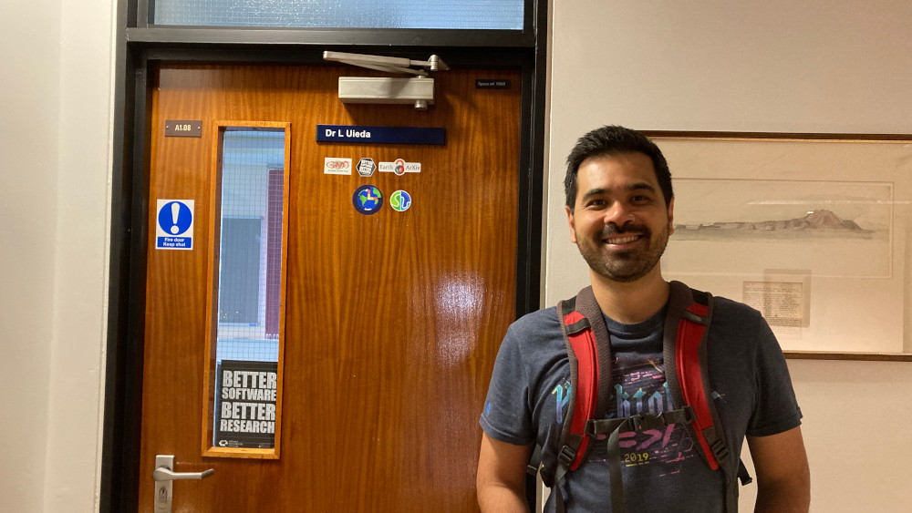 Photo of me smiling in front of my office door at the University of Liverpool. The door has 5 stickers and a name plate reading 'Dr L Uieda'.